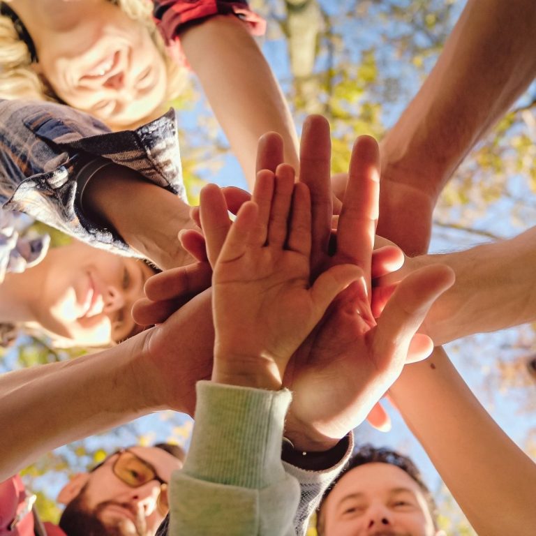 Team Eine Vielzahl an Händen ist in die Mitte gestreckt, darüber sind glückliche Gesichter zu sehen